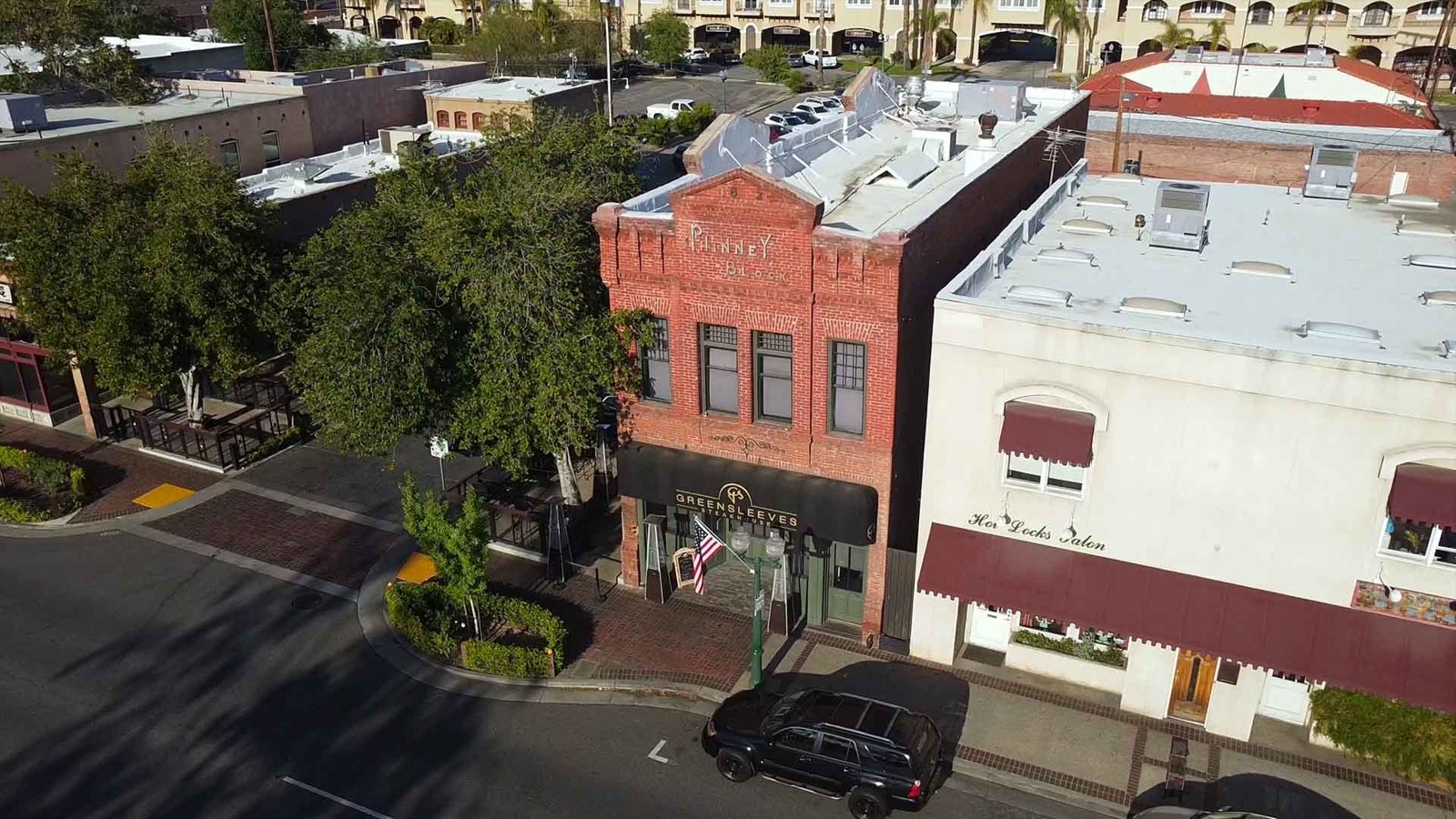 The corner from above, as the restaurant's own site opens: the brick Pinney Block building with the Greensleeves Steakhouse awning over the entrance, an American flag and patio heaters at the door, on a downtown Redlands street. Source: greensleevessteakhouse.com, the opening image of the home page, captured September 25, 2026.